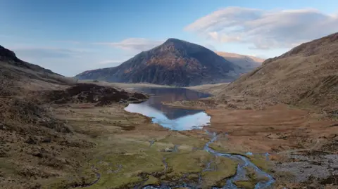 A view over Llyn Idwal lake with Pen yr Ole Wen mountain in the distance, at Snowdonia National Park. The green valley is large and wide, with the mountain in the horizon and blue sky with clouds behind. 