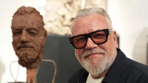 PA Media Ray Winstone is pictured close up with wearing large dark glasses smiling beside a clay sculpture of his bearded face.