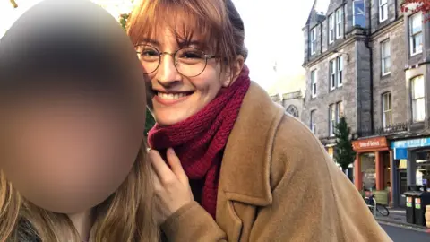 A young woman with auburn hair, large glasses, a cashmere coat and large red scarf smiles on a high street, next to another woman whose face is blurred