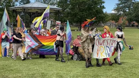 A line of people in drag and fancy dress costumes hold up rainbow flags and a banner saying Oswestry Pride as they walk across grass in a park, with a bandstand behind.