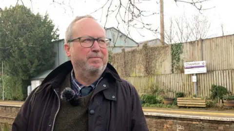 A man with grey hair and bear, wearing a black jacket and olive jumper standing in front of a train platform which is bordered by wooden planters and green shrubs 