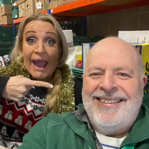 Stroud District Foodbank A close-up of Daisy May Cooper in a warehouse by shelving wearing a Christmas snowman jumper and tinsel scarf, pointing with an open mouth to a male worker who is smiling to camera. He has a green jumper and a short white beard. 