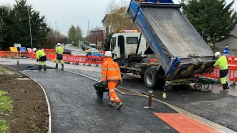 Wiltshire Council People in high-vis jackets doing work on a road. There is a large van and men in hard hats and red fences blocking off traffic.