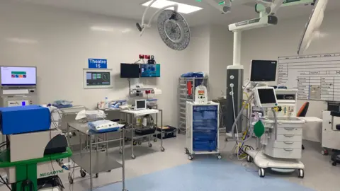 A surgical theatre - it's a large room with white walls and a grey floor. There are lots of hospital machines with screens, as well as trolleys on wheels with drawers of equipment. A sign on the wall reads 'Theatre 15' in white writing on a blue background.