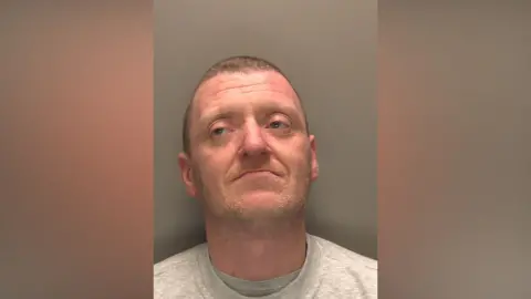 Custody photograph of Patrick Joseph Wiggins he has short brown hair and beard and is wearing a grey top