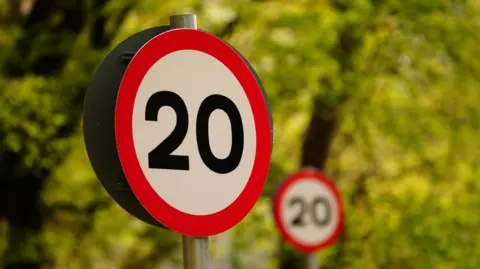A 20mph road sign stands before greenery, with another 20mph road sign in the distance.