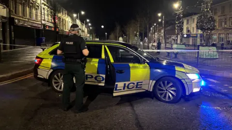 An officer has their back turned to the camera as they stand beside a police vehicle in Lurgan town centre. There is a large cordon.