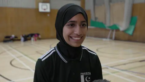 Haleema smiles widely to the left of the camera. She is wearing a black hijab and a black polo shirt with the Pakistan team logo on the top left. She is standing on a covered netball court with floor markings, the net and a hoop on the wall visible in the blurred background behind her