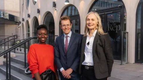 Kris Askey Three people stand outside the Birmingham Rep theatre: a woman in a red jumper, a man in a blue suit and glasses, and a blonde woman with a grey suit jacket