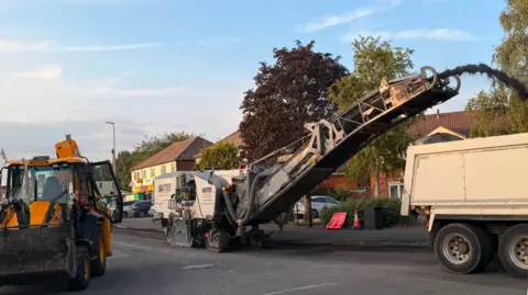 A yellow mechanical digger on a road alongside a beige truck and another machine pouring a black material into the back of it