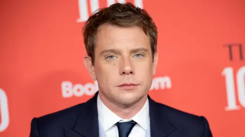 Jonathan Anderson stands in front of a large, red wall and looks at the camera. He has short hair, a navy suit jacket, a white collared shirt and a navy tie.