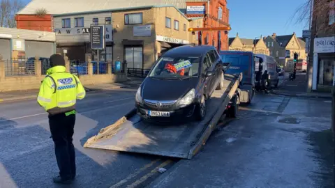 A car being winched onto a low loade with a police office in hi-vis vest standing guard on the roadside 