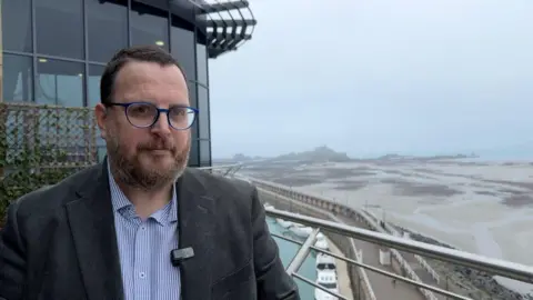 Chris Parker is wearing a blue pinstripe shirt, a grey suit jacket and glasses, in front of Jersey Port, with sand and boats in the background.