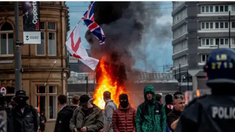 Getty Images Crowds gather in front of a burning building, with riot police visible in the corner of the image