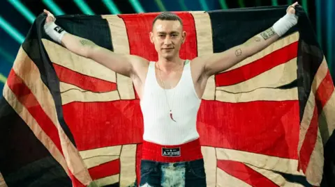 Getty Images Olly Alexander en Eurovisión en 2024. Olly sostiene la bandera de Union Jack sobre sus hombros mientras usa una camiseta de tanque blanca y pantalones cortos rojos visibles sobre sus jeans de mezclilla