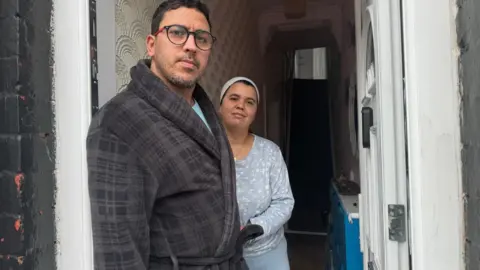 Jonny Humphries/BBC Mohammed Lamaoufi, 42, who has black hair, light stubble, and is wearing a grey cheque patterned dressing gown and glasses, stands in the doorway of a terraced house next to Khadeja Daoud, 34, who wears a light blue top and leggings