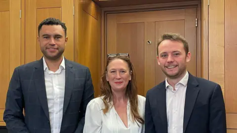 Supplied Two men in suits standing either side of a woman