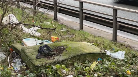 Mattress covered in moss and surrounded by other waste dumped on the side of a road