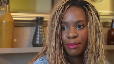 Muriel is seen sitting indoors in front of decorative bottles, looking slightly to one side.