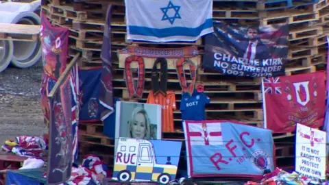 Wooden pallets stacked on top of each other. Draped across them are several flags, banners and scarves. There is also a large image of Michelle O'Neill. 