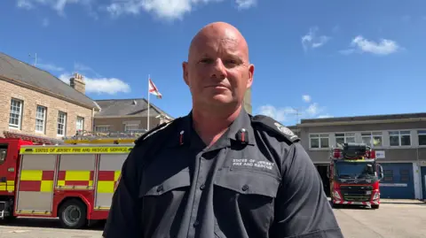 A fire officer in his uniform standing in front of fire engines at a fire station.