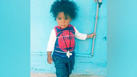 A young child in dark blue trousers and white top under a blue vest.