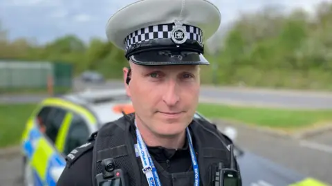 Jim is wearing a white police cap and standing in front of a police car