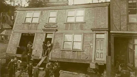 West Cheshire Museums/Lion Salt Works archive A building called Castle Chambers has sunk backwards, leaving a hole in the ground beneath it. A number of men are around it, with three men standing in the doorways of the building looking out. 