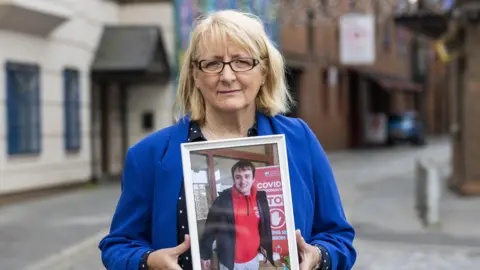 Dawn Jones has blonde hair and wears black glasses. She is dressed in a blue coat and a dark blouse with white polka dots. She is holding a framed photo of her son Timothy, who is wearing a red polo shirt and a dark jacket.