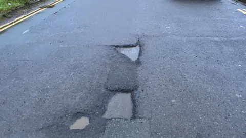 A road with three potholes in the centre. The potholes are filled with water. There are double yellow lines either side of the picture. There is a grass verge to the left.