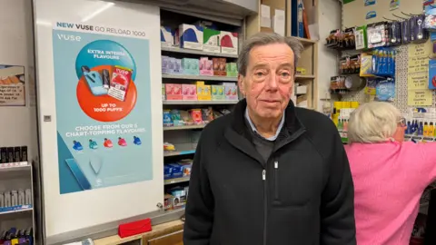 An elderly man with grey hair looking at the camera. There is a cabinet of vapes over his left hand shoulder.