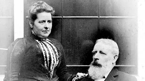 Charles Wells Josephine and Charles Wells are shown in an old black and white photo. Charles is sitting down to the right and Josephine is standing to the left. She has her hair up and is wearing a Victorian bodice. Charles has grey hair and a long beard. 