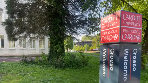 A tall rectangular sign stands on grass beside trees and a path. The sign reads “Cardiff University / Prifysgol Caerdydd” in white text on a red background, with “Croeso / Welcome” printed vertically below on a dark panel. Behind the sign is a light‑coloured stone building partly obscured by trees and foliage.