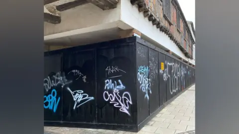 Stephanie Northen A black hoarding with graffiti in different colours scribbled on it can be seen, with a Tudor building's frontage above it