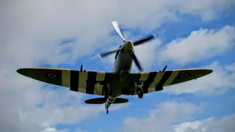 ARRAN GORDON A spitfire which is light green and has black and white stripes on the wings flying against a blue sky with patches of white cloud. The propeller at the front of the plane is blurred as it is moving.