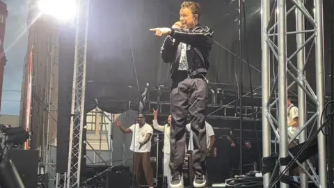 BBC / Zoe Toase Olly Murs is singing and jumping in the air on stage, with his band in the background. He is smiling and pointing at the crowd.