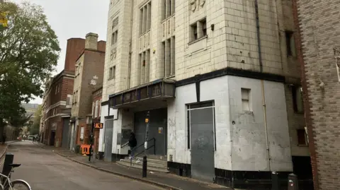 A three-storey art deco cinema with tall, narrow windows on the upper stories, and an entrance door flanked by stairwell entrances on both sides of the ground floor. There is wooded hoarding above the door where the name would have been. There is a narrow road passing the cinema.