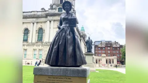A bronze statue of a women in a large frock. She is wearing a bonnet on her head and holding in front of her a piece of paper with a depiction of someone on their knees praying. Behind the statue is the lawn of Belfast City Hall.
