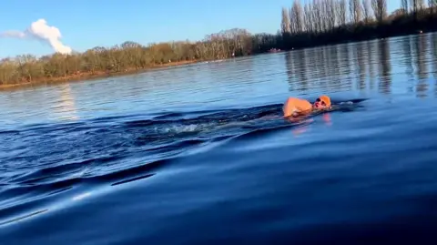 A woman wearing a pair of googles and an orange swimming hat swimming in a lake with trees at the water's edge.