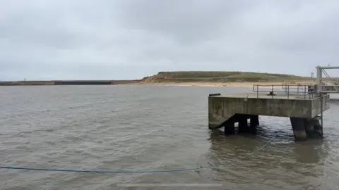 Andrew Turner/BBC A concrete "dolphin" - a structure used to moor ships alongside while they load or discharge cargo, usually stone or sand minerals. In the far distance is the undeveloped southern terminal, which is made of sand, much of which is covered in sand. In the foreground is sea water in the outer harbour.