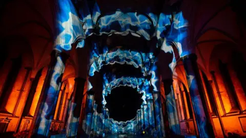 Tom Gregory The nave of a huge cathedral with coloured swirling lights all over in blue and orange