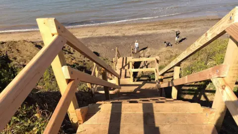 Steep wooden staircase down to a beach. Several people are at the bottom on the sandy beach.