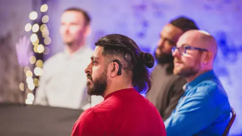 Becky Bailey/LumoTV Four men are sat around a table. One man is in the centre and he has a cochlear implant and is wearing a red t-shirt. 