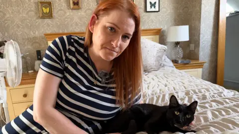 Kelly Burns is sat on a bed with a black cat. Kelly is wearing a blue and white striped dress. She has medium-length ginger hair. She is holding the black cat's face with her left hand.