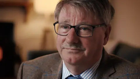 Firecrest Films Head shot of a man with brown/grey hair combed in a side shed, glasses and a moustache. He has a serious expression on his face and is wearing a brown checked blazer, a white shirt with blue stripes and a blue patterned tie.