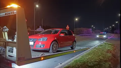 A red car is half way up a platform leading from the road onto a recovery truck. A police car with its lights flashing is a short distance behind on the same road. Grass verge is on one side and the central reservation barrier is on the other.