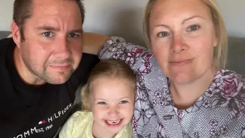 Family sat together man on the left wearing a black t shirt, woman on the right wearing a purple flower print top and daughter in the middle smiling