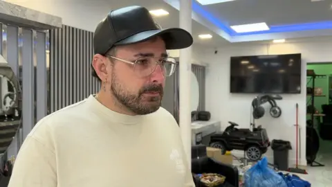 Mariano is wearing a black cap and clear-framed glasses stands inside a barbershop or salon. The person has facial hair and multiple ear piercings and is dressed in a light-colored T-shirt. In the background, there is a wall-mounted TV, salon chairs, equipment, blue plastic bags, and a small toy car placed near the floor, with bright overhead lighting and blue accent lights on the ceiling.