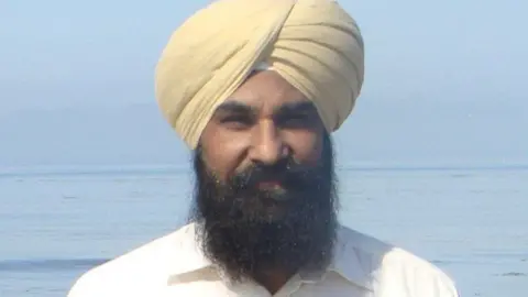 Family Anakh Singh, wearing a turban and standing on the shore with the sea and small waves behind him.