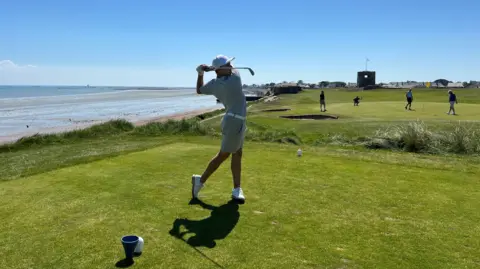 Josef has swung his golf club on a green with others playing the background and to the left there is a beach and seaside. Josef is wearing white shoes, cream shorts and t-shirt and a white cap.